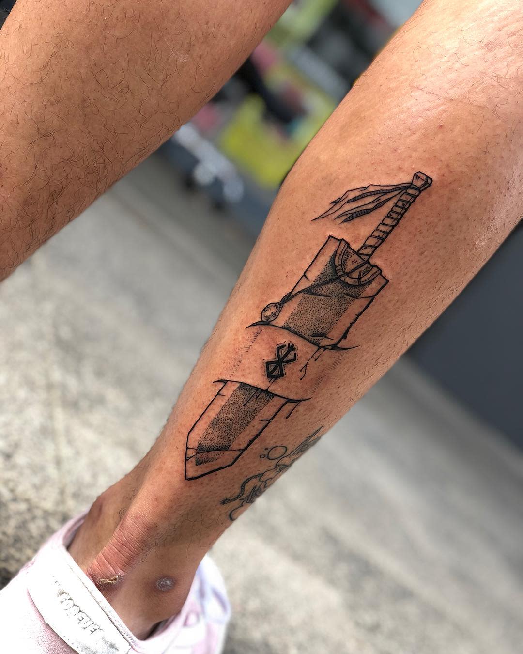 Linework tattoo of Guts' Dragon Slayer sword from Berserk on the calf, featuring the Brand of Sacrifice on the blade and a flowing ribbon tied to the hilt